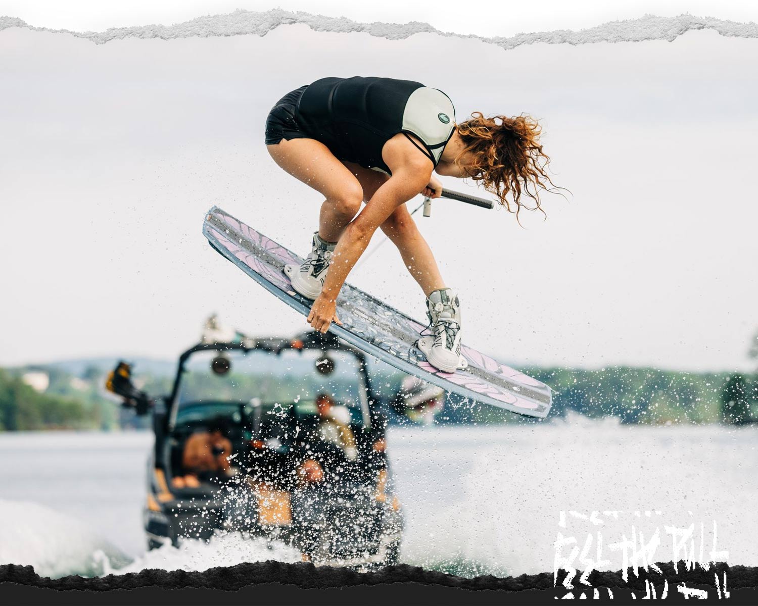 Woman in tan vest grabs wakeboard midair while being towed behind a boat.