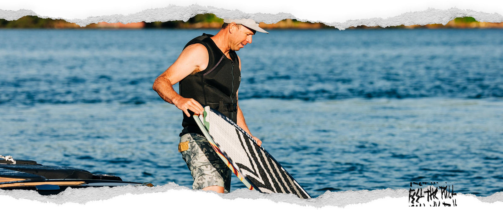 Man in black Watson CGA vest with gray trim steps off boat while holding a white wakesurf board.