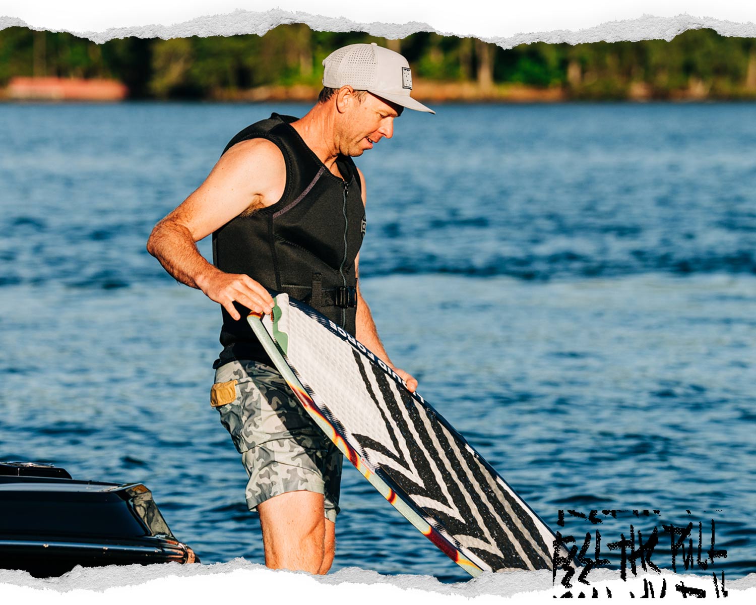 Man in black Watson CGA vest with gray trim steps off boat while holding a white wakesurf board.