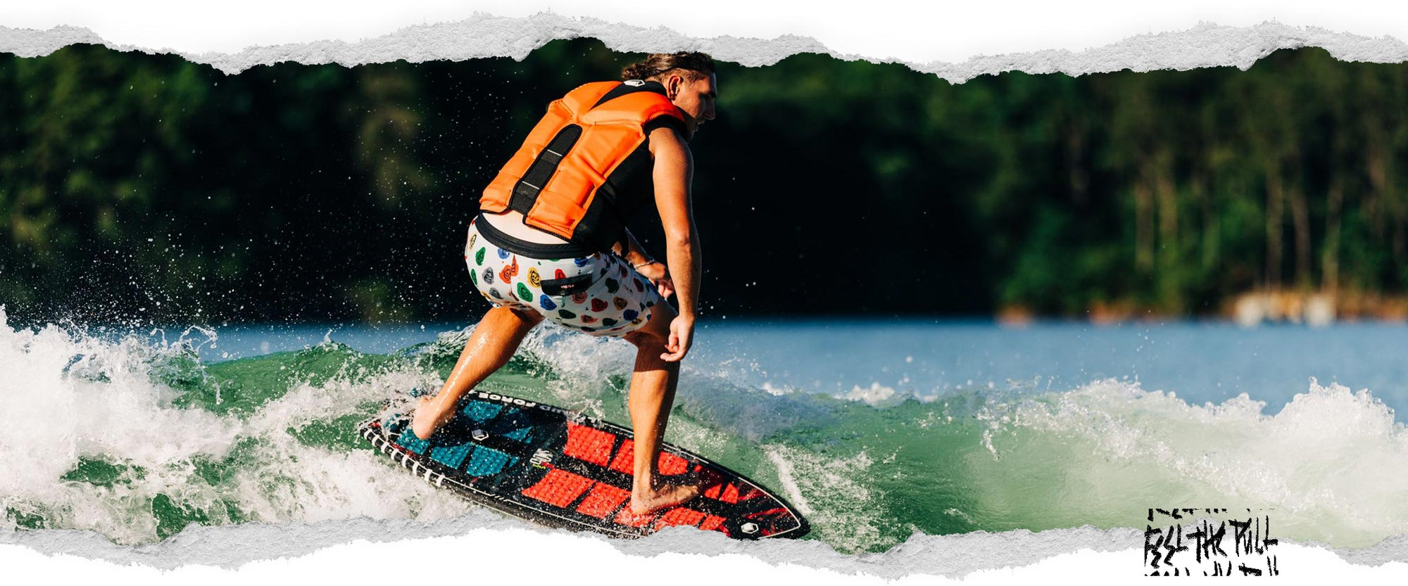 Rider surfs a wake while wearing a bright orange and black Watson CGA vest and printed shorts.