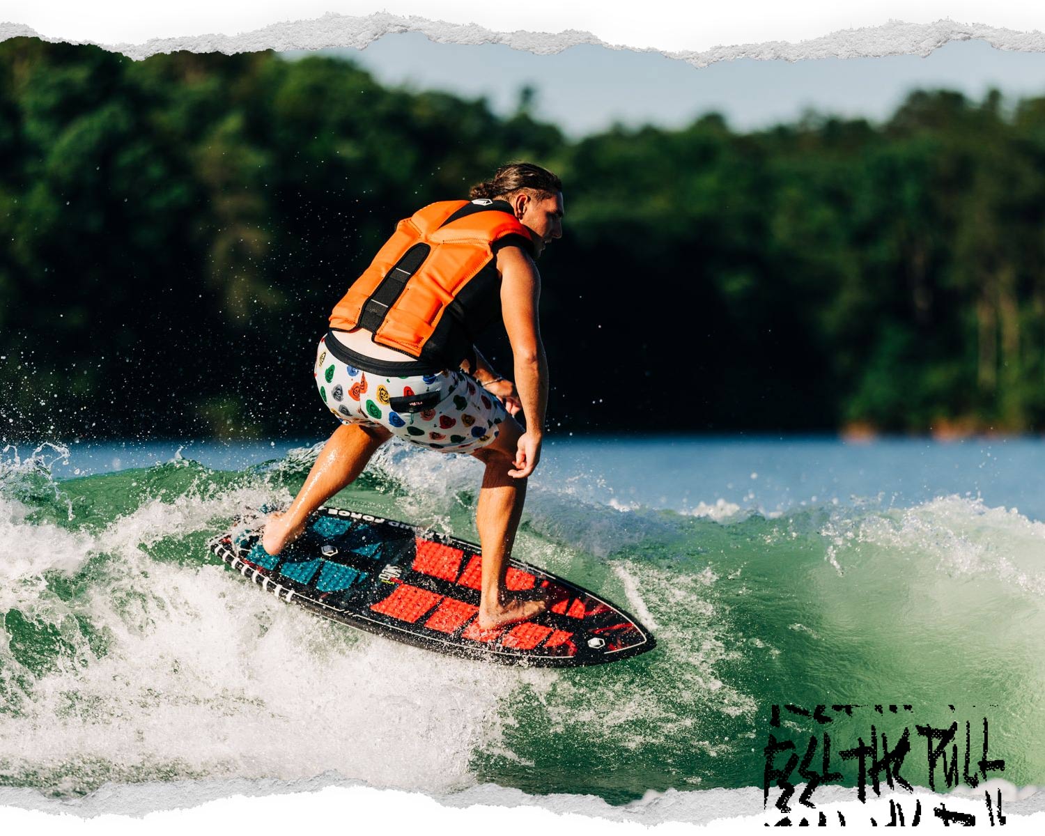 Rider surfs a wake while wearing a bright orange and black Watson CGA vest and printed shorts.