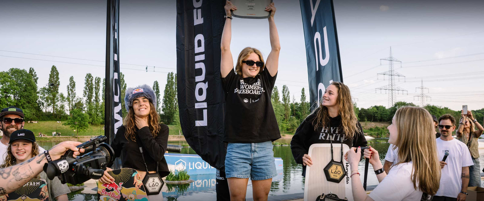 Woman in a black Women's Wakeboarding Tee raises a trophy on a wakeboarding podium.