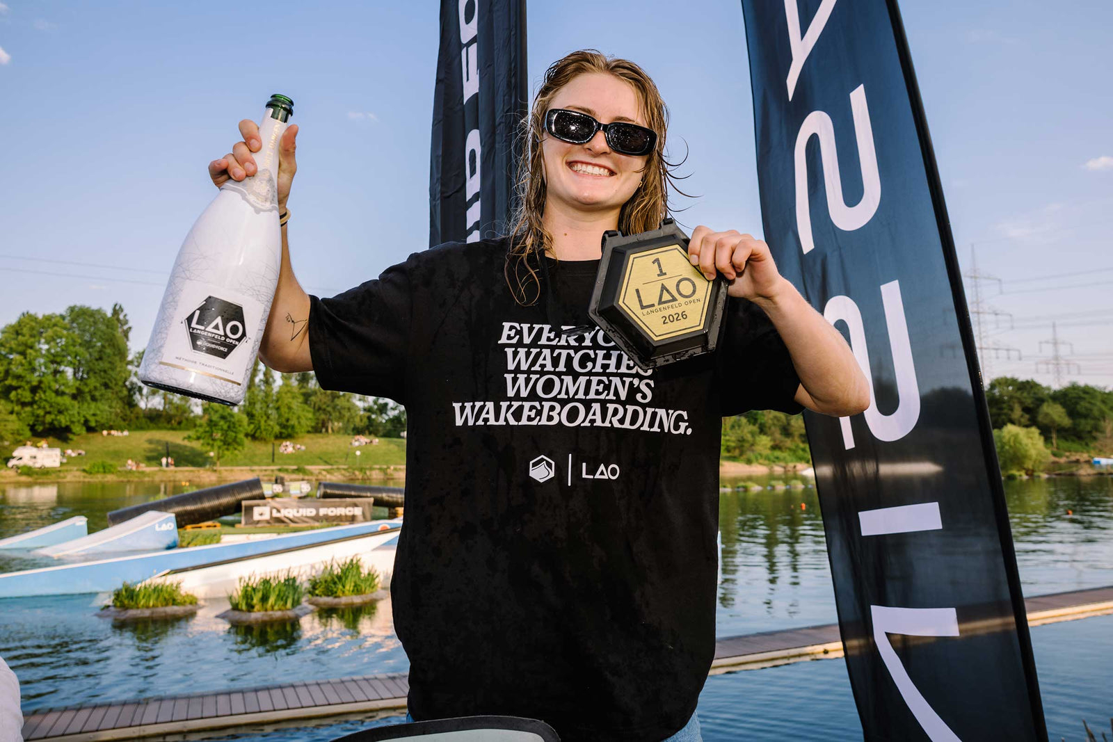 Woman wearing a black Women's Wakeboarding Tee holds a trophy and bottle by the water.