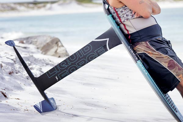 Person leaning against a LiquidForce foil board in the sand with water in the background. The wake foil features a blue thin LiquidForce logo. He is wearing black and brown shorts with a colorful life vest.