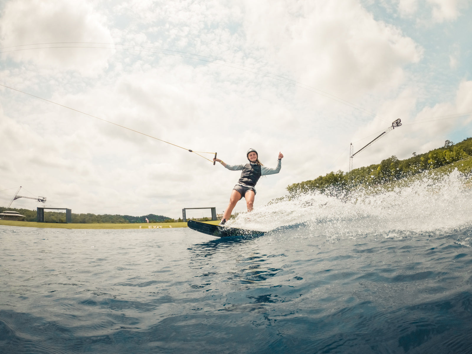 Wakeboarder rides across the lake on a cable system, smiling with one hand off the rope.